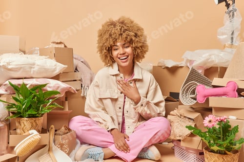 Preview: Happy beautiful woman with curly hair moves in new place sits crossed legs on floor surrounded by ca