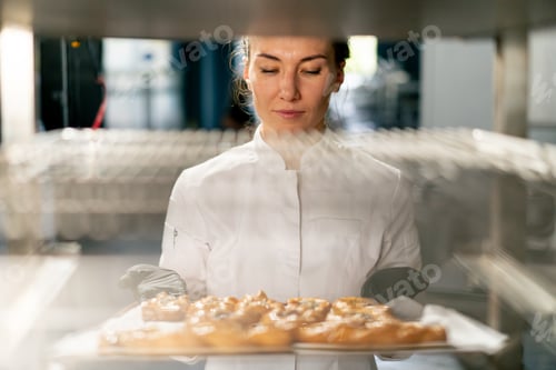 Preview: A female baker wearing gloves takes out a baking sheet with freshly baked hot bread from the oven