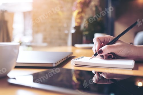 Preview: woman's hand writing on a blank notebook with laptop , tablet and coffee cup on wooden table