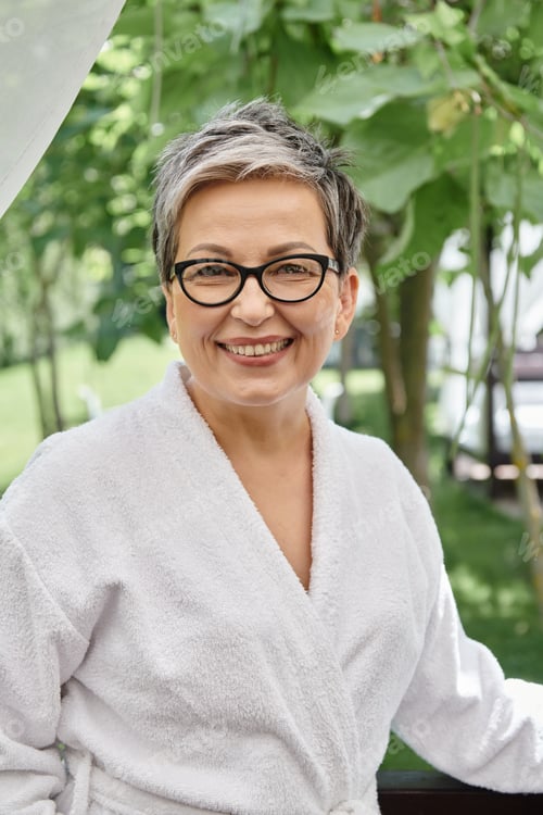 Preview: happy middle aged woman in glasses and white robe looking at camera during wellness retreat