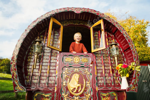 Preview: A boy leaning out of the open shutters at the rear of a bow top gypsy caravan.