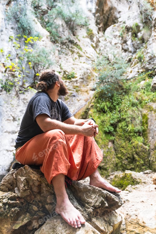 Preview: Caucasian man with beard and long hair looking at the sky while sitting on a rock next waterfall