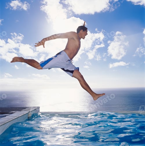 Preview: Man in swimming trunks jumping into a swimming pool by the ocean, sky and clouds.