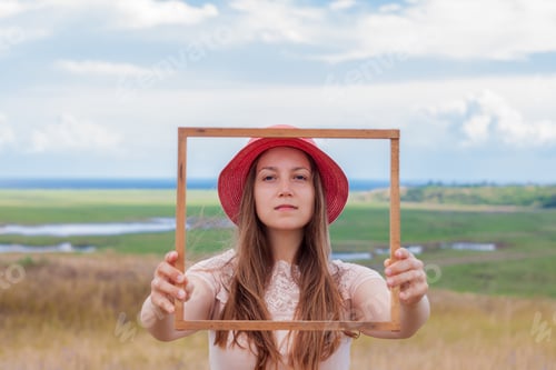 Preview: Girl in pink hat holds wooden picture frame on nature landscape background portrait Creative idea