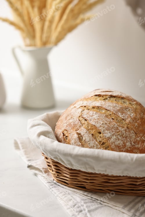 Preview: Wicker bread basket with freshly baked loaf on white marble table in kitchen, closeup