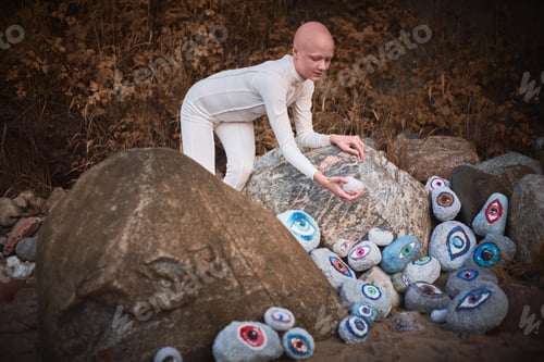 Preview: Young hairless girl with alopecia in white futuristic costume looking at surreal lot of rocks eyes