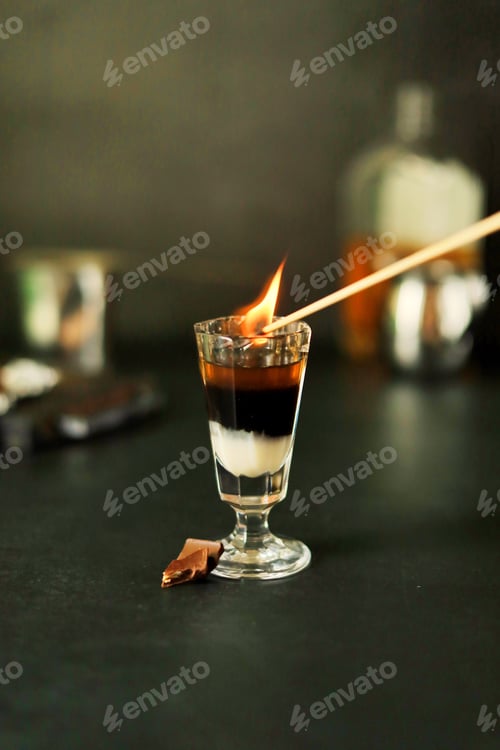 Preview: A thin stick ignites a three-layer cocktail shot in a glass, and it burns with a flame