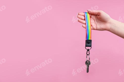 Preview: Woman holding key with keychain in LGBT colors on pink background, closeup. Space for text