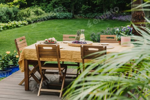Preview: Dining table covered with orange tablecloth standing on wooden terrace in green garden