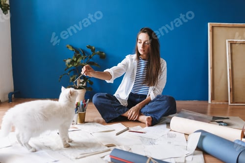 Preview: Thoughtful girl sitting on floor with drawings dreamily looking