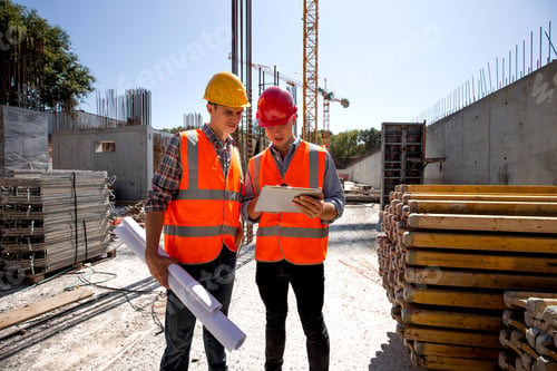 Preview: Civil architect and construction manager dressed in orange work vests and helmets discuss a building
