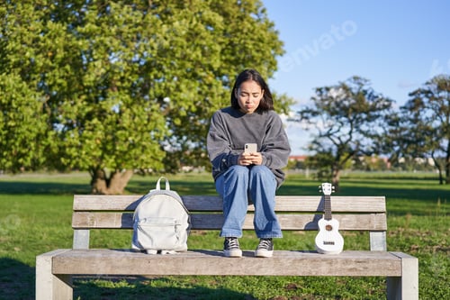 Preview: Beautiful brunette girl on bench in park, sitting with ukulele and backpack, holding smartphone