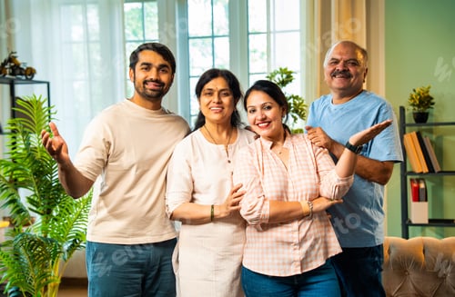 Preview: Smiling at camera, Indian family of four standing together in cozy and modern living room