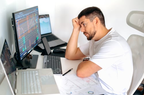 Preview: Focused male trader sitting at desk at home office monitoring stock market looking at monitors