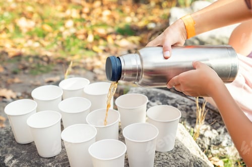 Preview: Close-up top above view woman hand hold steel thermos pouring hot tea many white paper cup group