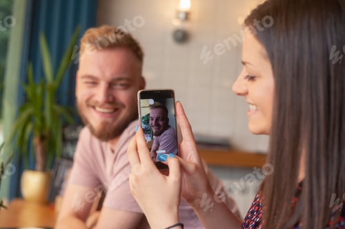 Preview: One Girl Talking A Picture Of A Young Man, On A Smartphone. In A Cafe.