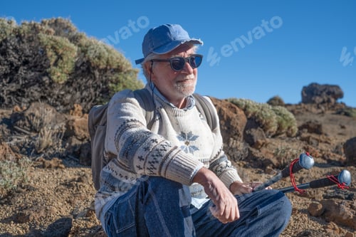Preview: Active happy senior man with backpack in outdoor excursion stops to rest looking the rocky landscape