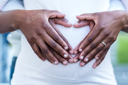Preview: Mid section of couple forming heart shape on belly while standing in park