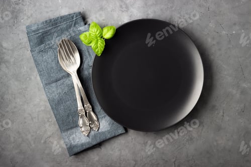 Preview: Black plate, cutlery, napkin and basil leaves over gray concrete background.