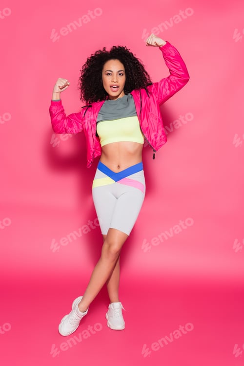 Preview: full length of sporty brunette african american woman showing muscles while posing on pink