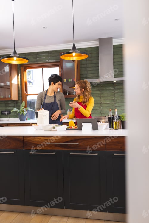 Preview: Happy biracial lesbian couple using tablet and cooking in kitchen, copy space