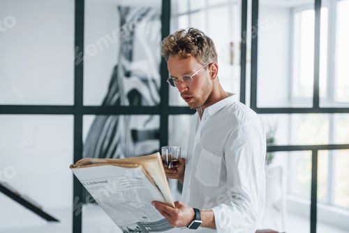 Preview: Reads newspaper. Caucasian young guy in elegant white shirt indoors at home
