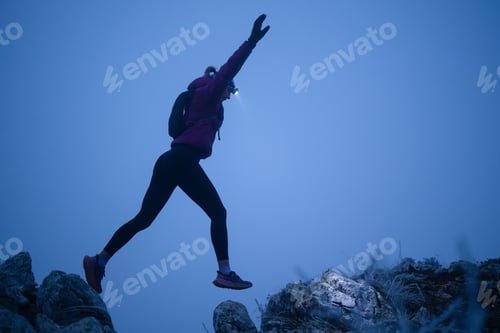Preview: silhouette of a fitness woman trail runner jumping over a cliff and running to a rocky mountain top