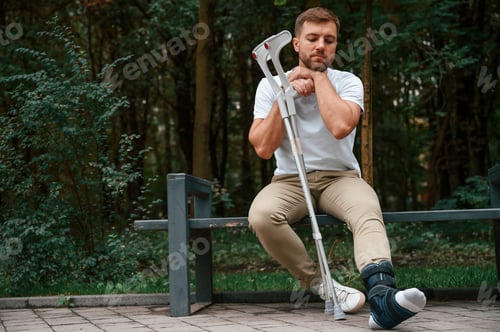 Preview: Sitting, feeling bored and sad. Man with crutches is in the park outdoors. Having leg injury