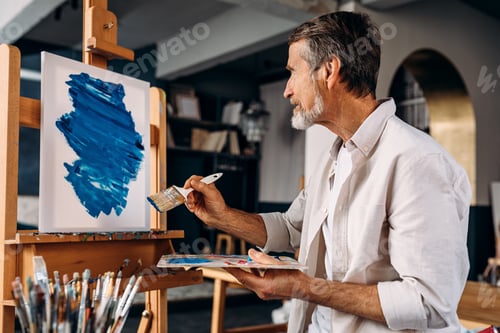 Preview: Bearded artist sitting in front of easel and painting abstract picture with large paintbrush