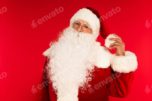 Preview: Bearded santa claus in eyeglasses touching hat isolated on red
