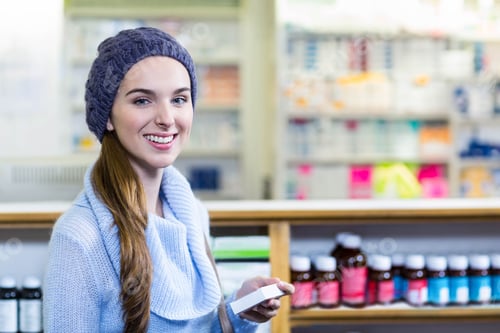 Preview: Teenage girl standing at pharmacy counter holding medicine box and smiling in beanie and sweater