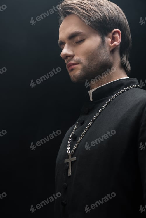 Preview: young catholic priest standing in darkness with closed eyes isolated on black