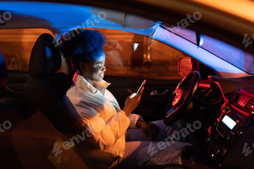 Preview: Happy black girl sit in car uploading social media. Young african american female smartphone addict