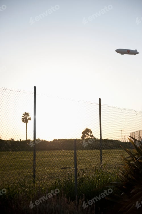 Preview: Blimp flying over park with palm trees and wire fence in foreground.