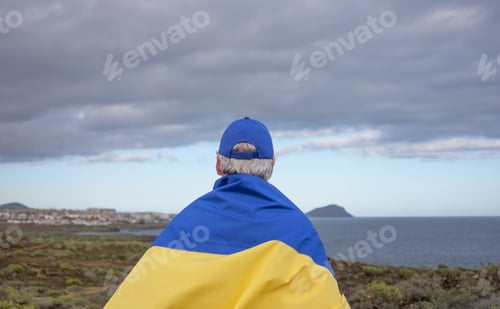 Preview: Rear view of a senior caucasian man looking out to sea with the Ukrainian flag on his back