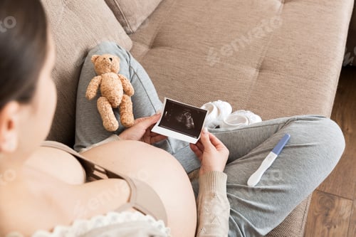 Preview: Pregnant Woman sitting on bed and Looking At Ultrasound Scan photo of her Baby.