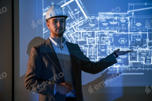 Preview: Male architect in hard hat is standing against projector with building plan on it