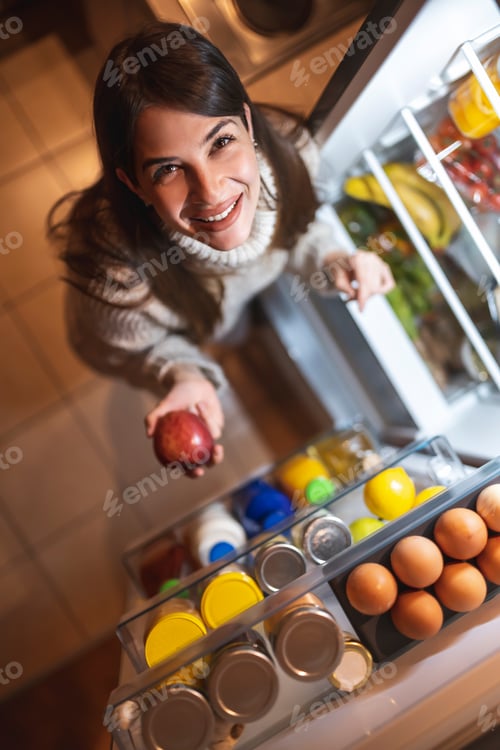 Preview: Woman holding an apple in front of an opened fridge