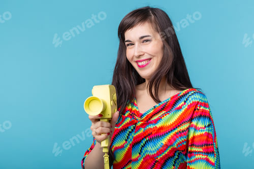 Preview: Beautiful positive young brunette woman holding in her hands a model of a yellow camcorder posing on