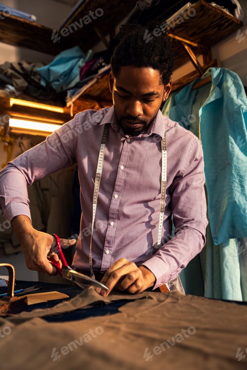 Preview: Male fashion tailor working on the suit design in his workshop cutting fabric with scissors.