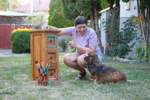 Preview: Woman painting a wooden furniture while sitting with her dog in the garden. DIY concept.