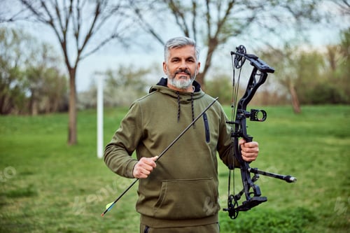 Preview: Portrait of a smiling senior bearded man in archery, holding bow and arrow.
