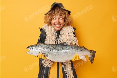 Preview: Horizontal shot of positive female angler holds big caught fish wears black raincoat and hat carries