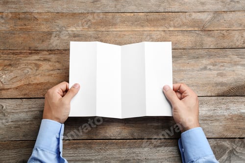 Preview: Young man holding blank brochure at wooden table, top view. Mock up for design