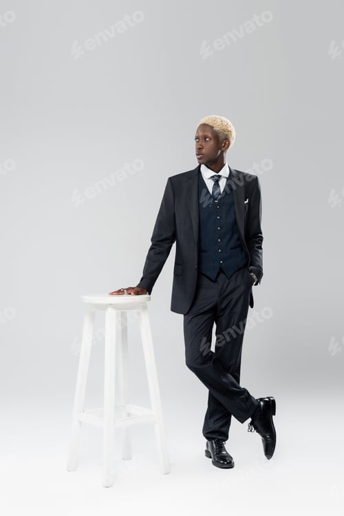 Preview: full length of blonde african american man in suit standing with hand in pocket near white chair on