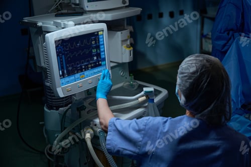 Preview: Young female assistant in gloves, mask and uniform pointing at display of medical equipment