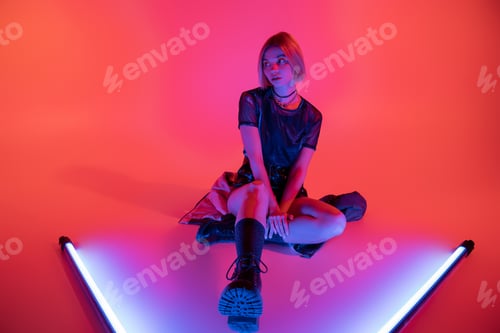 Preview: full length of woman in trendy boots sitting near purple neon lamps and looking away on coral