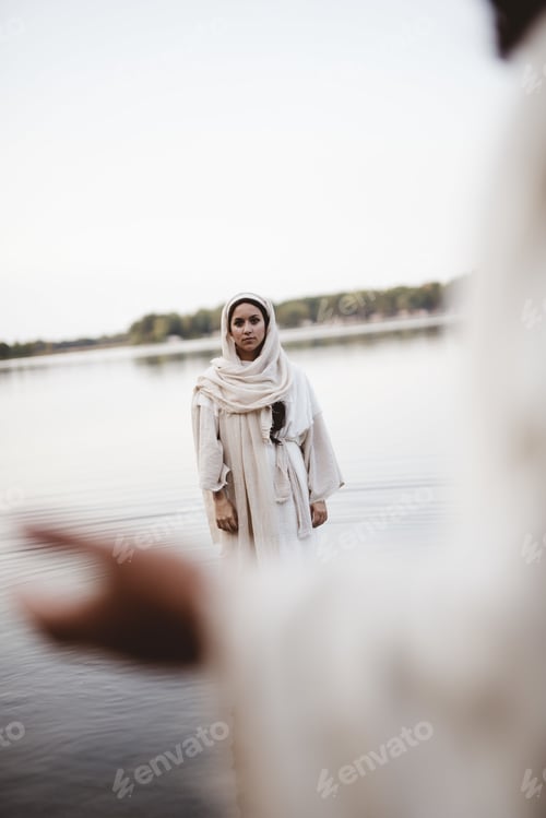 Preview: Vertical shot of a female wearing a biblical robe while standing in water looking at Jesus Christ