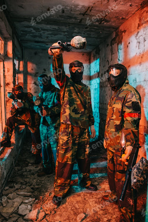 Preview: male paintball player aiming by gun at camera while his paintball team in uniform and protective