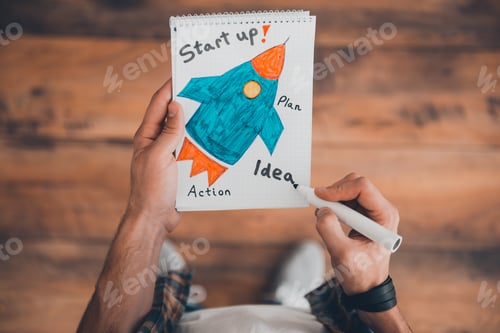 Preview: Planning his own start up. Top view of man drawing in note pad while standing on the wooden floor